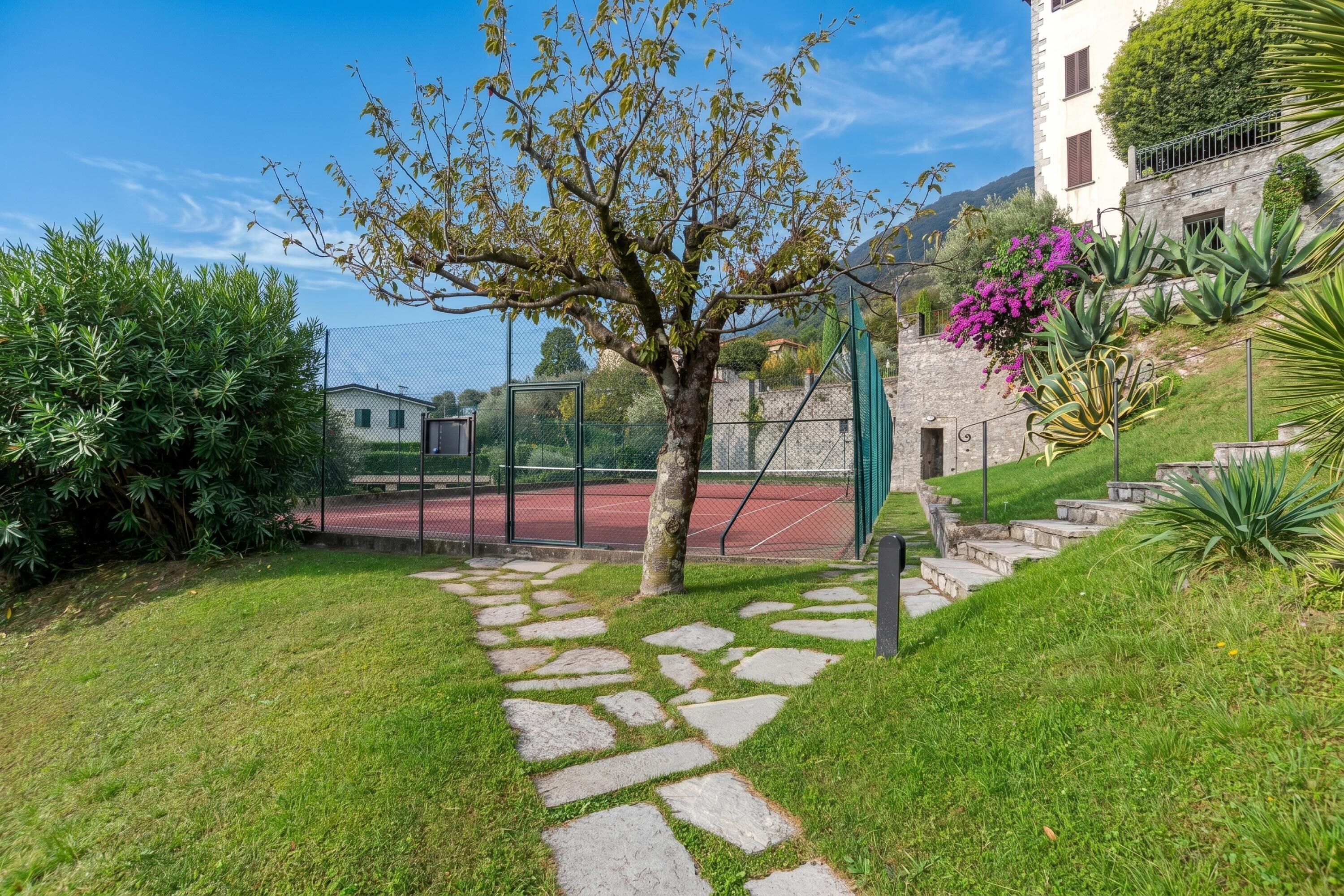 En stendækket sti der fører til en tennisbane, omgivet af grønt og en bygning med balkon.
