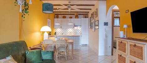 A living room with a sofa, a dining table with chairs, a kitchen in the background, and a TV on a cabinet.