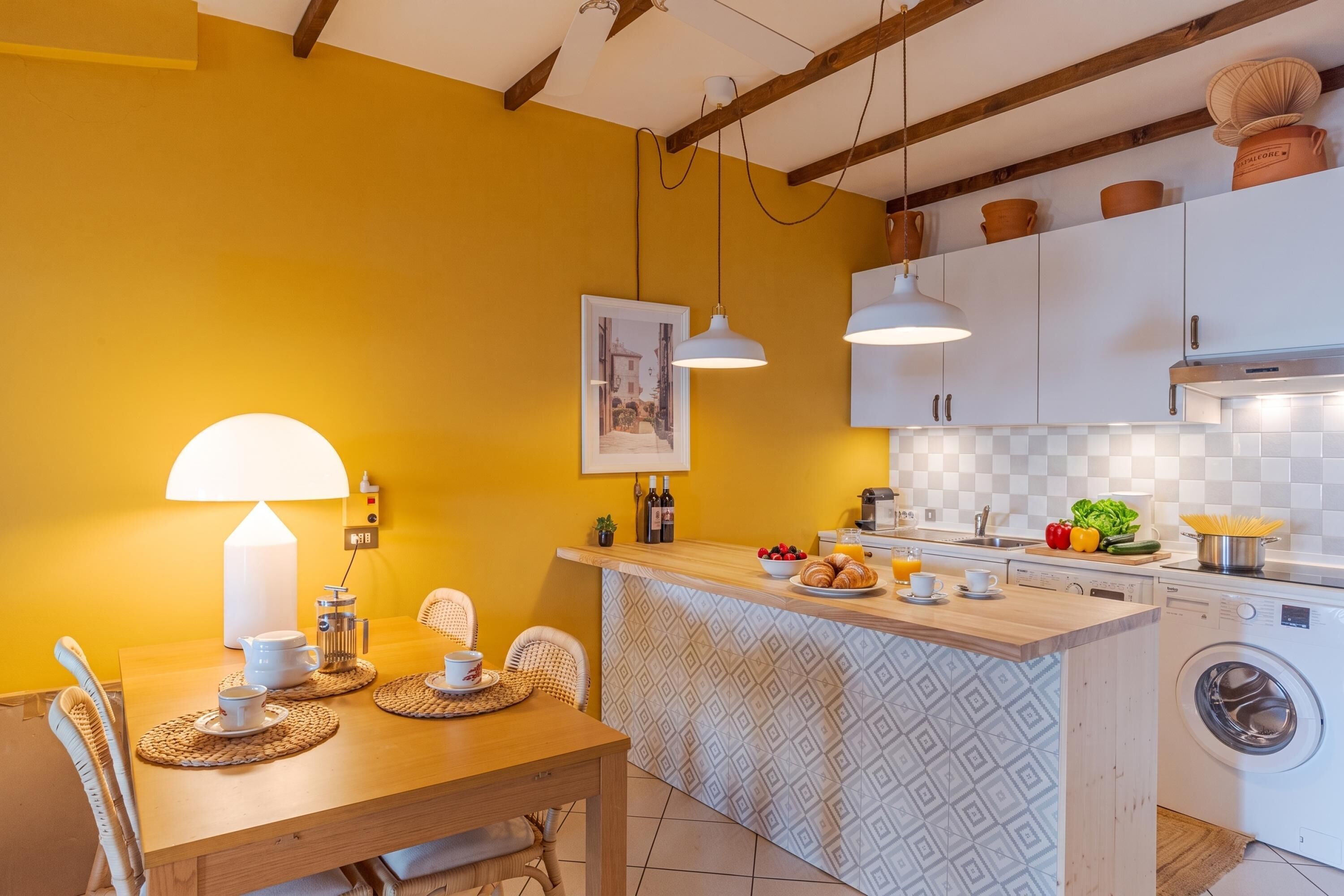 Una cocina con pared amarilla, mesa de madera, luces colgantes blancas y una lavadora.
