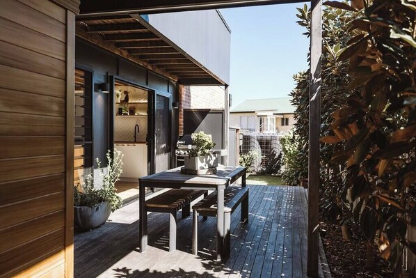 An outdoor patio with a table and benches, surrounded by plants and a wooden deck.