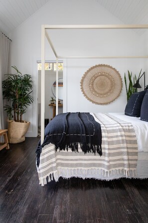 A bedroom with a bed, a woven wall hanging, a potted plant, and a wooden floor. - Soul of Gerringong Farmhouse (Gerringong)