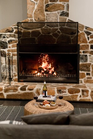 Una chimenea de piedra con fuego, una botella de vino, copas y una mesita con aperitivos.