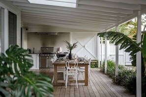 A covered outdoor dining area with a wooden table and chairs, a grill, and a kitchen in the background.
