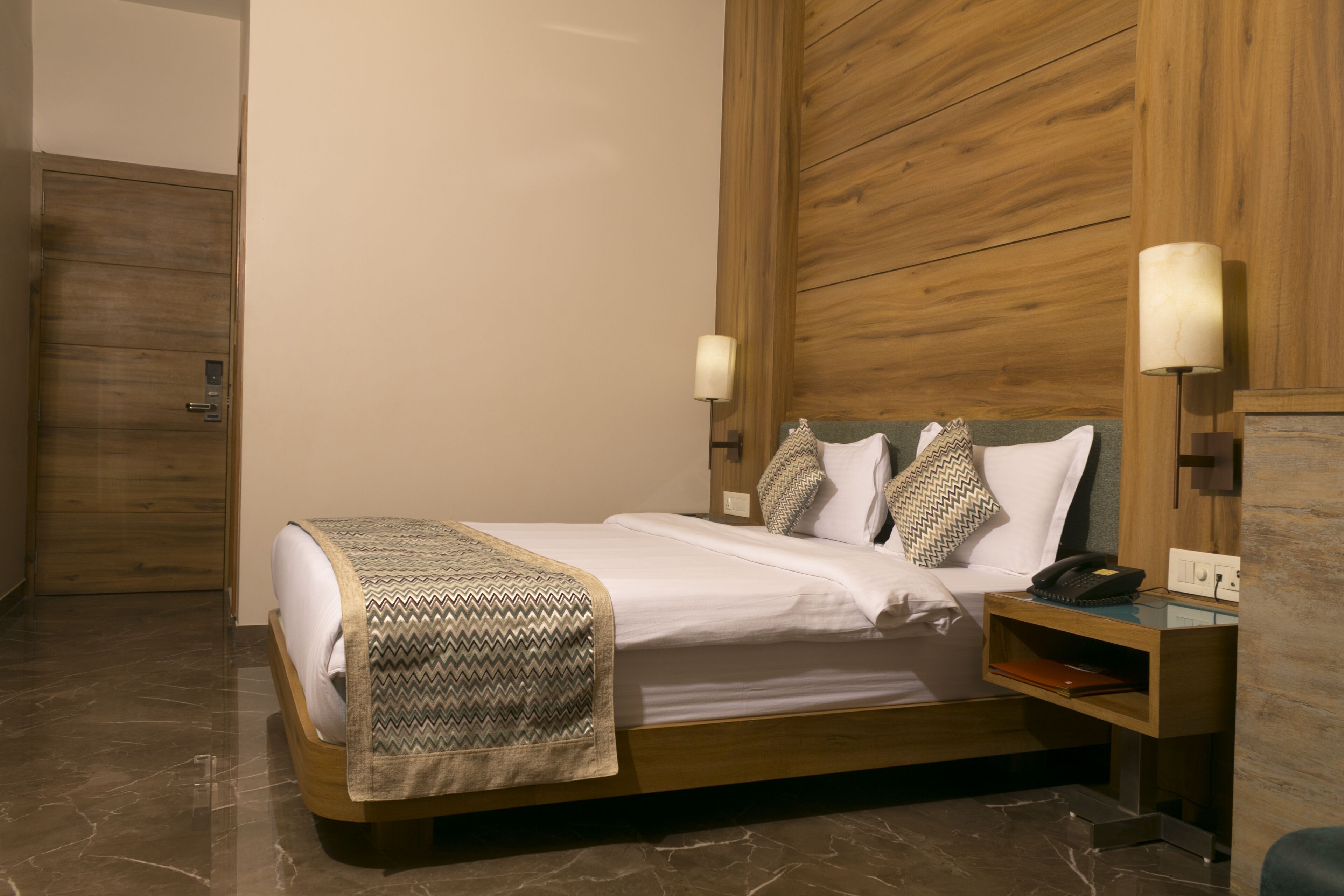 Una habitación de hotel moderna con un cabecero de madera, una mesita de noche con lámpara y suelo de mármol.