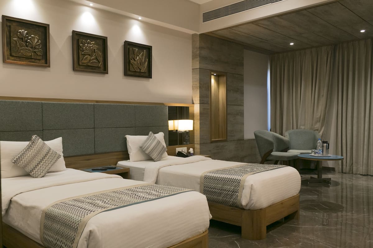 A hotel room with two beds, a desk, a chair, and a small table. There are wall-mounted lamps and framed artwork.