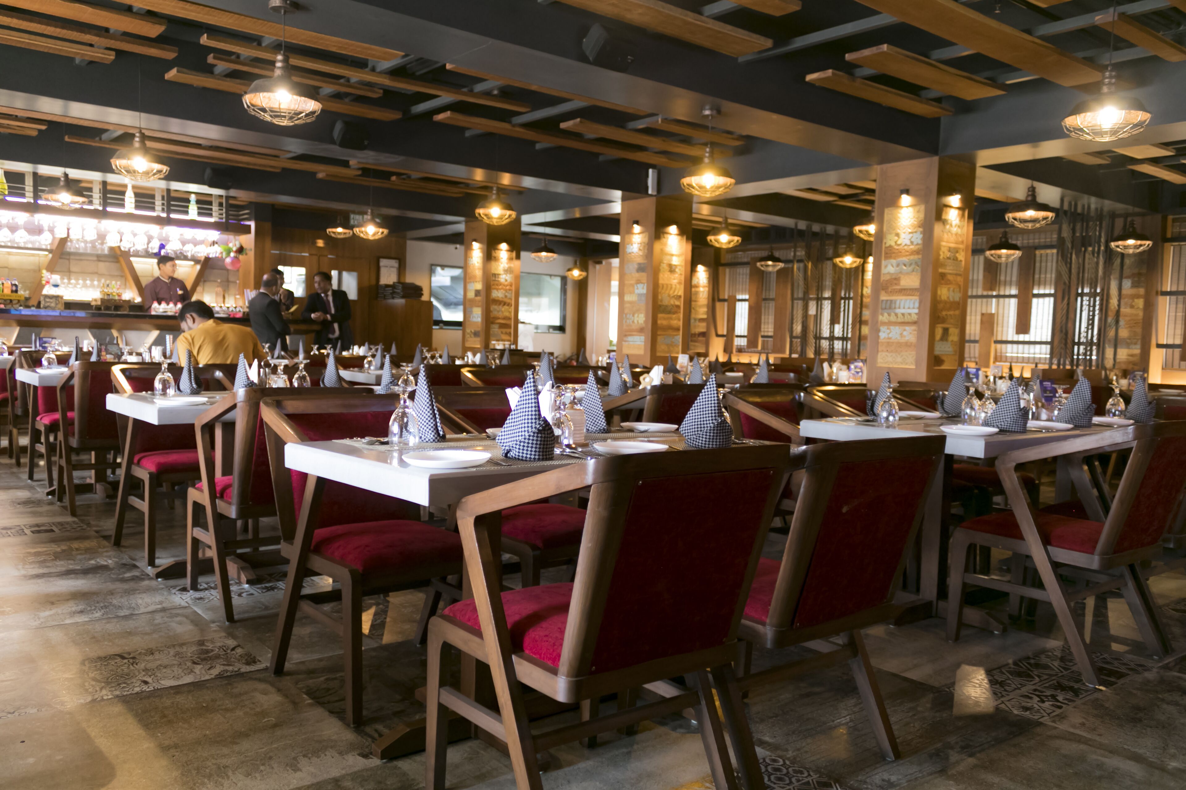 Interior de restaurante com vigas de madeira no teto, cadeiras estofadas vermelhas e mesas postas com toalhas brancas e guardanapos azuis.