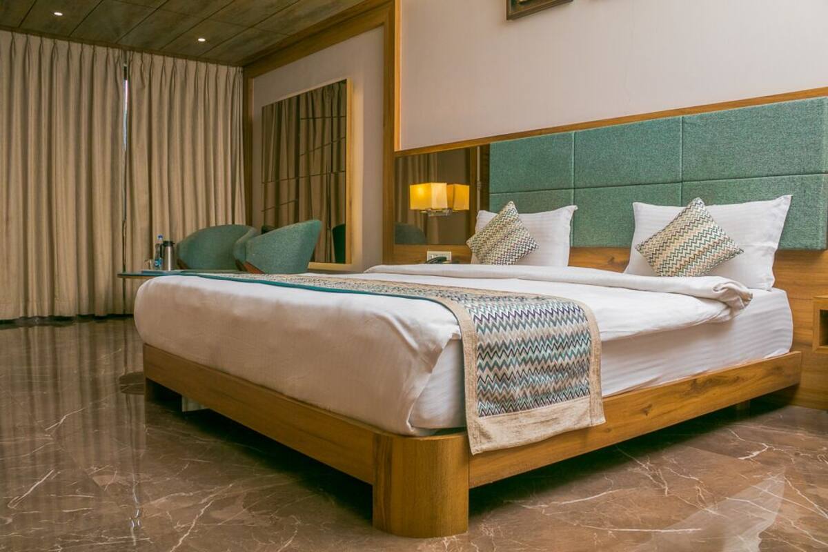 A hotel room with a large bed, a wooden headboard, a nightstand, a lamp, and a view of the outside through a window with curtains.