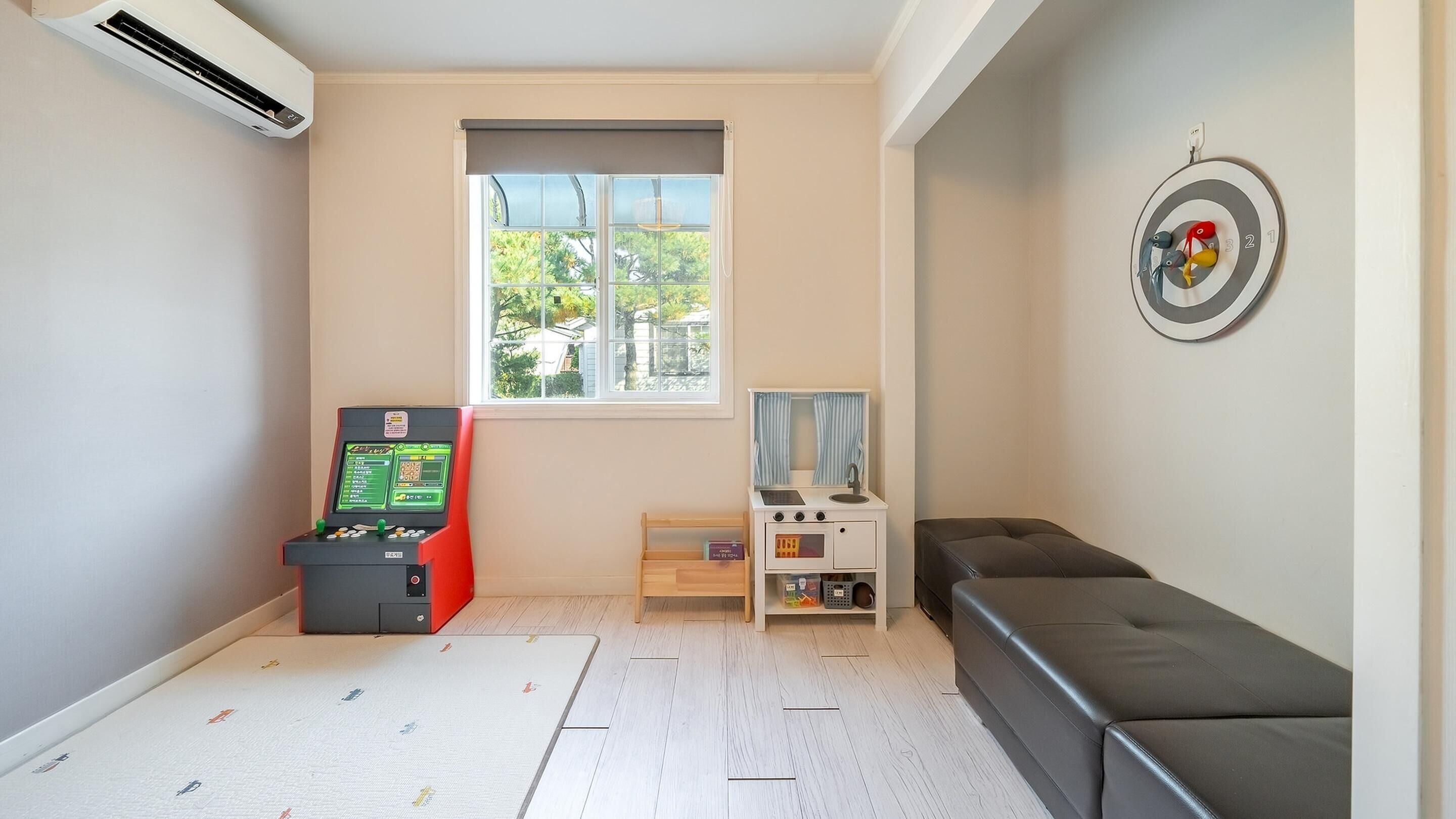A room with a window, a target on the wall, a sofa, a small kitchenette, and a gaming console.