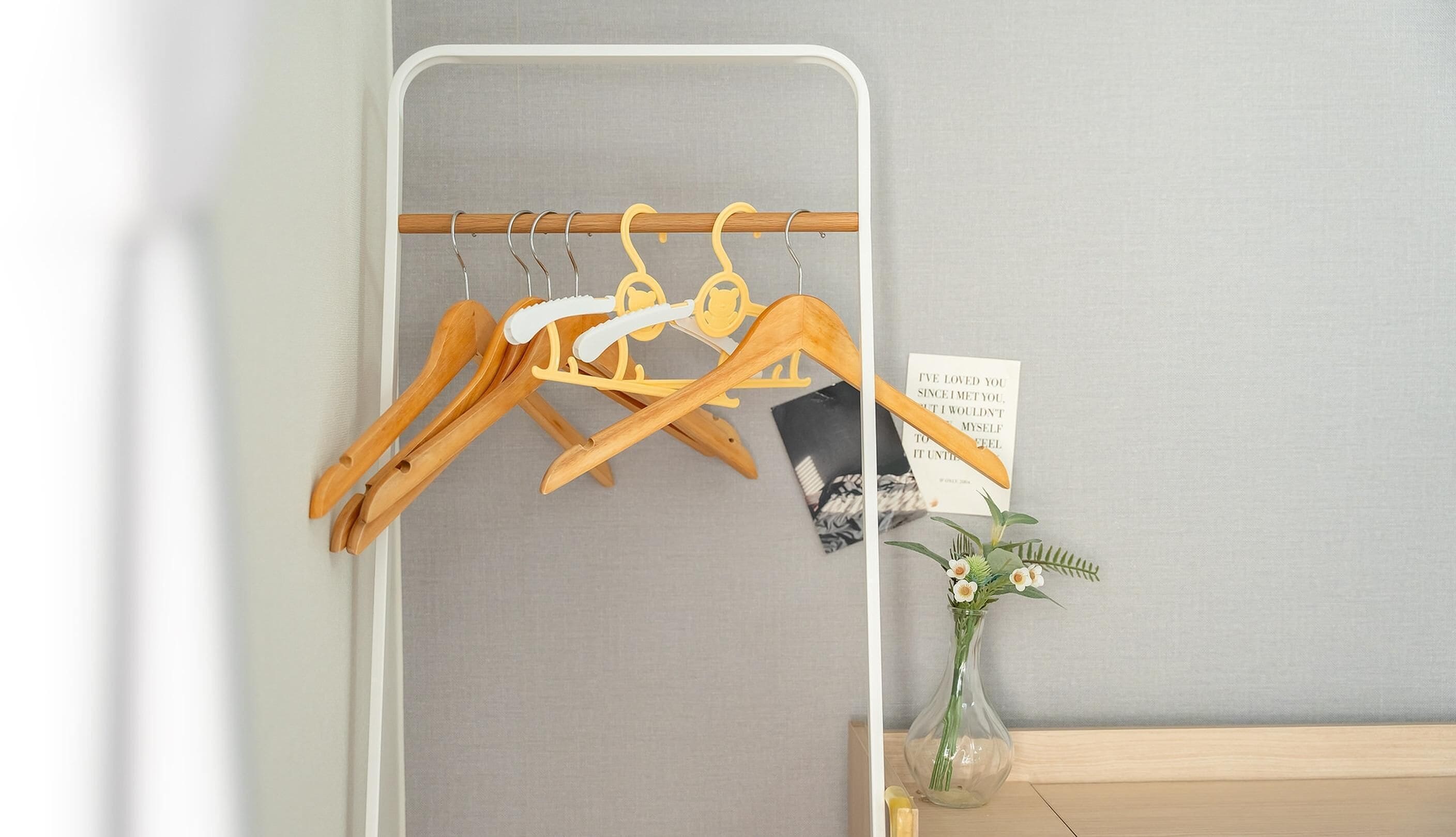A room with a wooden hanger rack against a grey wall, a small vase with flowers, and a framed quote on the wall.