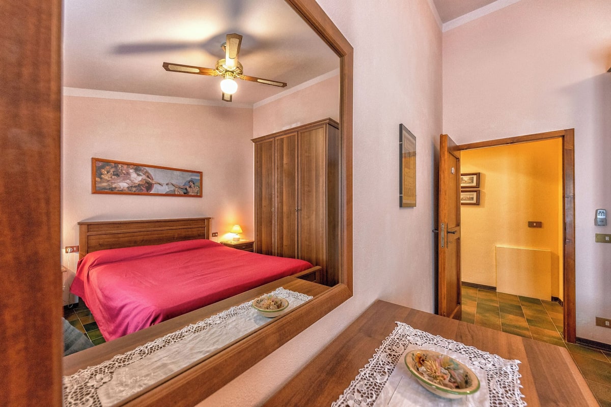 A bedroom with a bed, a wooden wardrobe, a ceiling fan, a painting, and a door leading to another room.