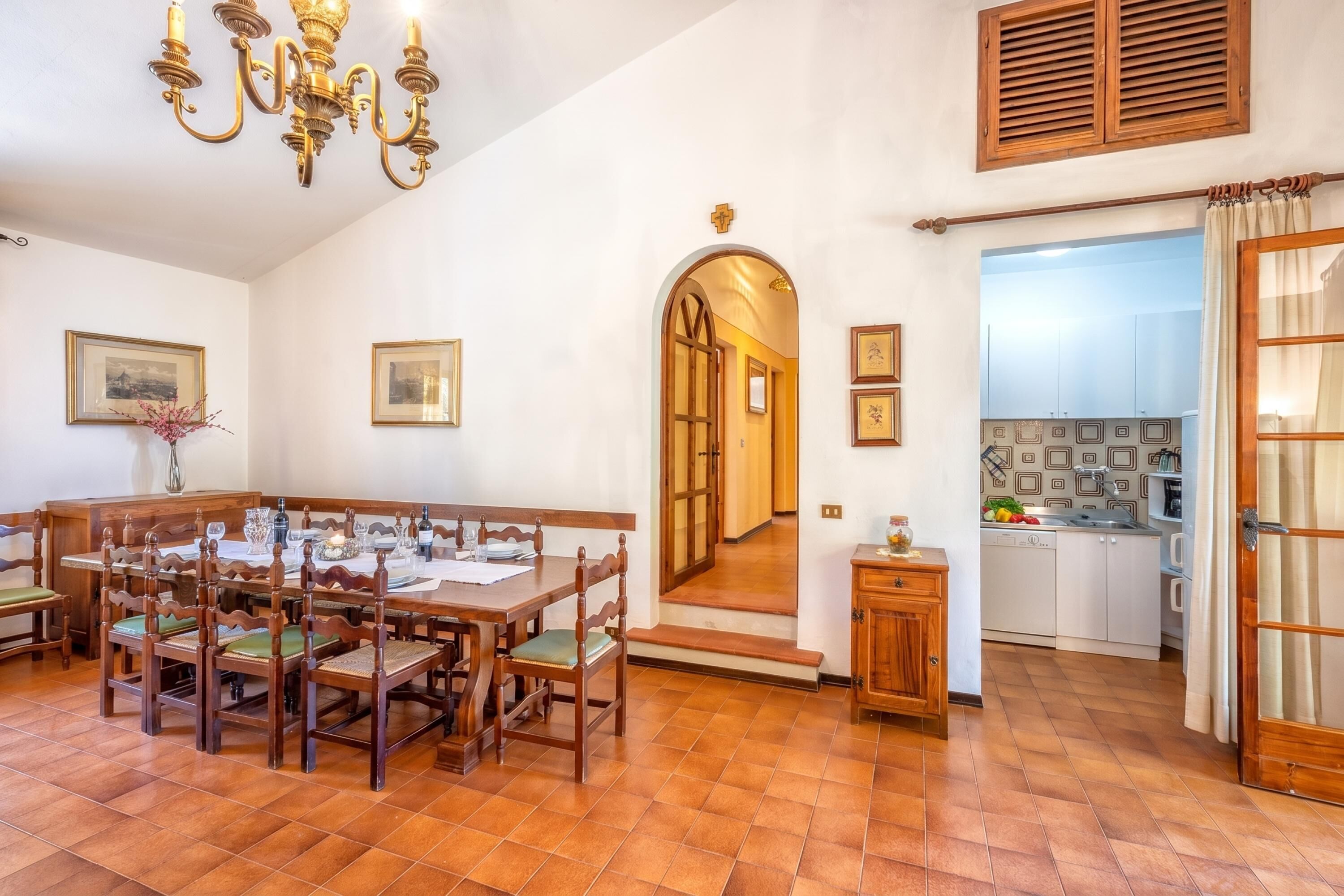 A dining area with a wooden table and chairs, a chandelier, and a kitchen visible through an archway.
