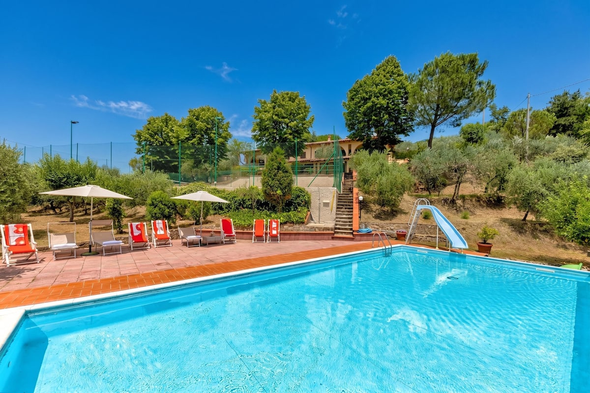 A swimming pool with a slide, surrounded by lounge chairs and umbrellas, with trees and a building in the background.