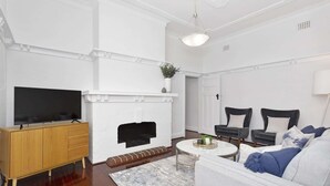 Una sala de estar con chimenea, un televisor sobre un mueble de madera, dos sillas negras, un sofá blanco y una mesa de centro redonda.