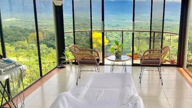 A room with a bed, two wicker chairs, a small table, and a keyboard. It has large windows offering a view of a green landscape. - Mountain Top Park Hotel (Quepos)