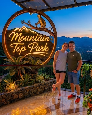 Ein Paar steht vor einem leuchtenden Neon-Schild mit der Aufschrift 'Mountain Top Park', das eine Berg- und Affenabbildung zeigt.