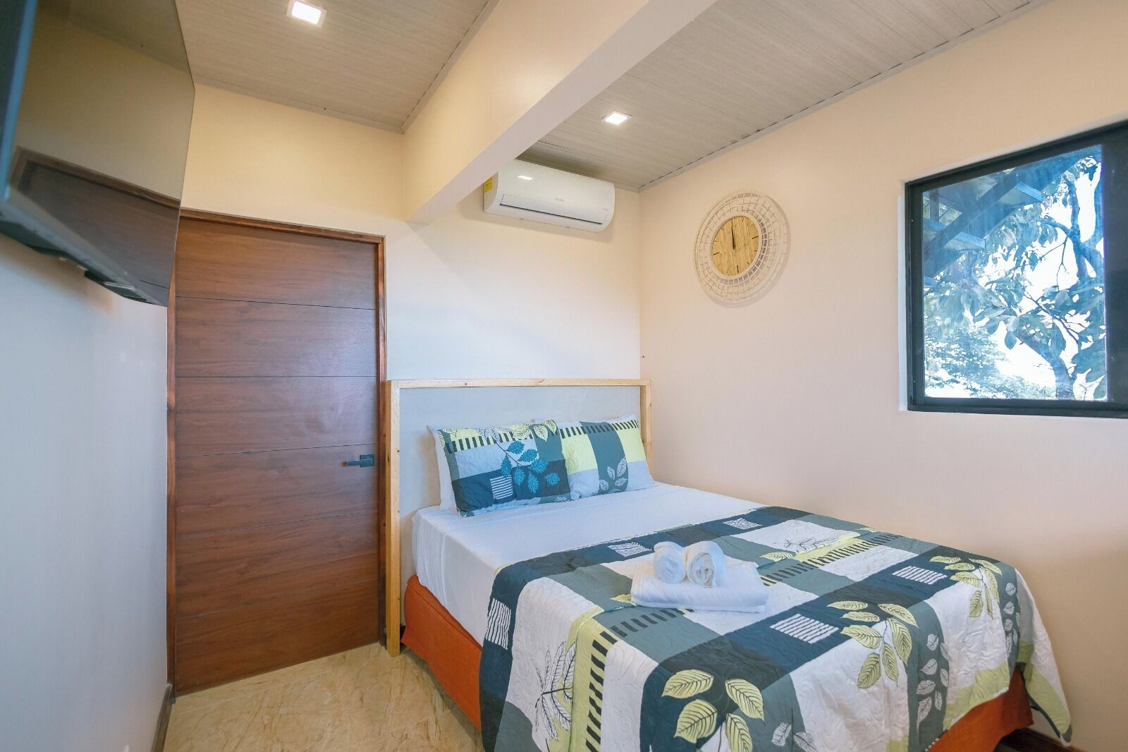 Une chambre avec une porte en bois, un lit recouvert d’une courtepointe, une horloge murale et une fenêtre donnant sur des arbres.