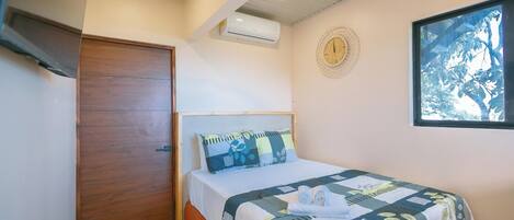 Un dormitorio con puerta de madera, cama con colcha, un reloj de pared y ventana con vistas a árboles.