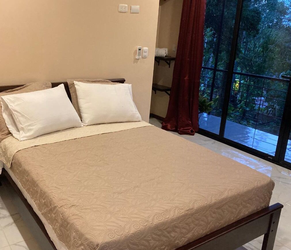 Een net opgemaakt bed met een beige sprei en twee kussens, een nachtkastje met een lamp, en een groot raam met uitzicht op bomen.
