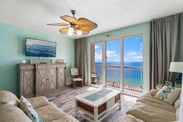 A living room with a view of the ocean, a ceiling fan, a television, and a dining area with a table and chairs.
