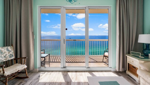 A room with a view of the ocean through a sliding glass door, a wooden rocking chair, a small table, and a lamp. - Seychelles 2103 (Panama City Beach)