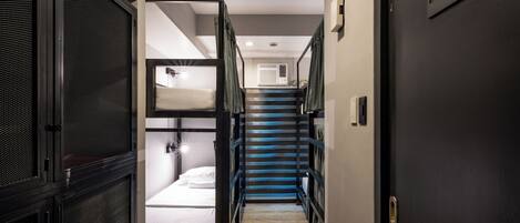 Un pasillo estrecho con literas a la izquierda, una escalera en el centro y el letrero de un baño a la derecha.