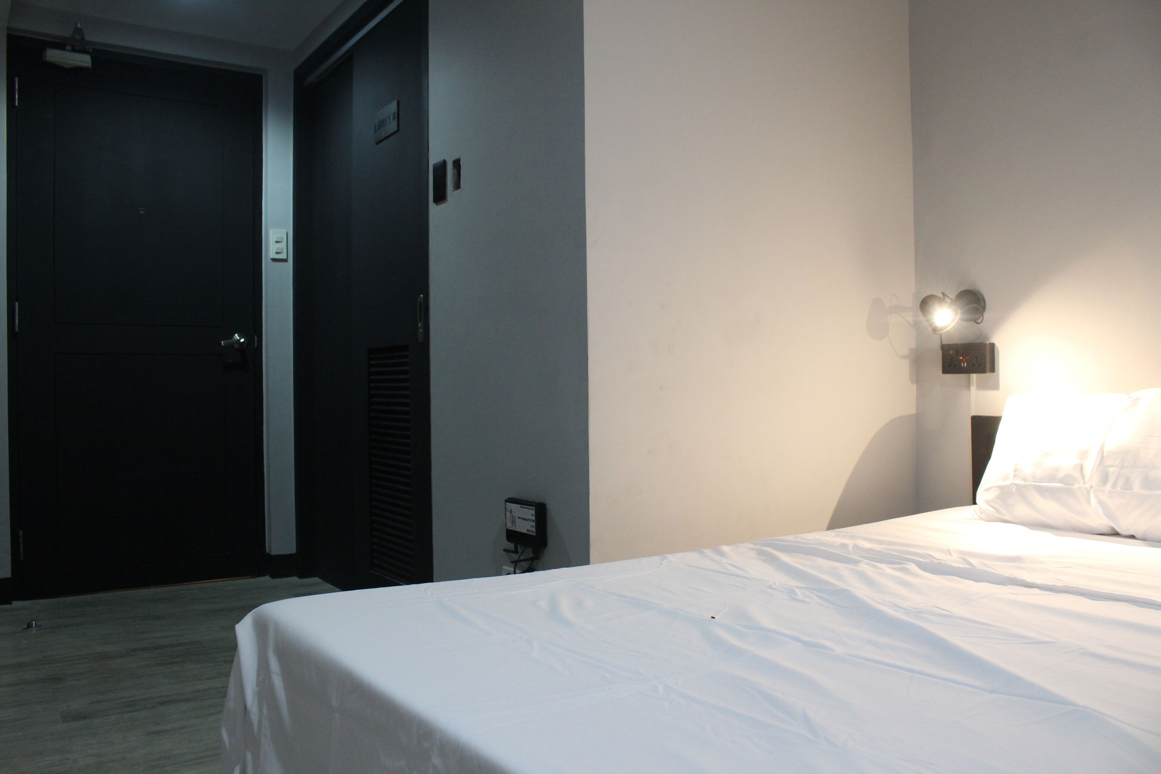 A hotel room with a bed, a door, a wall-mounted light, and a small electrical outlet.