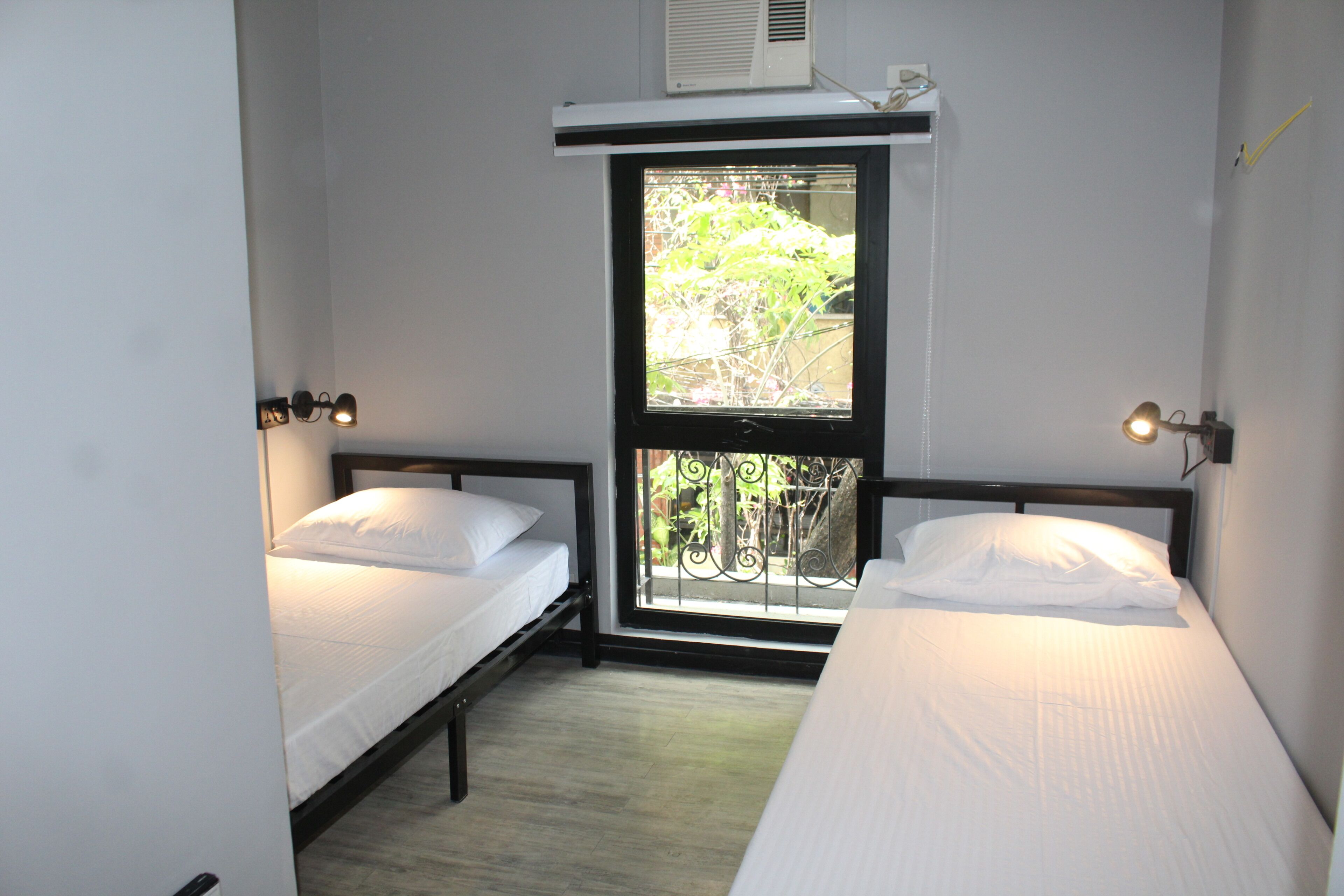 Une petite chambre propre, avec deux lits simples, une fenêtre donnant sur un paysage verdoyant et un ventilateur de plafond.