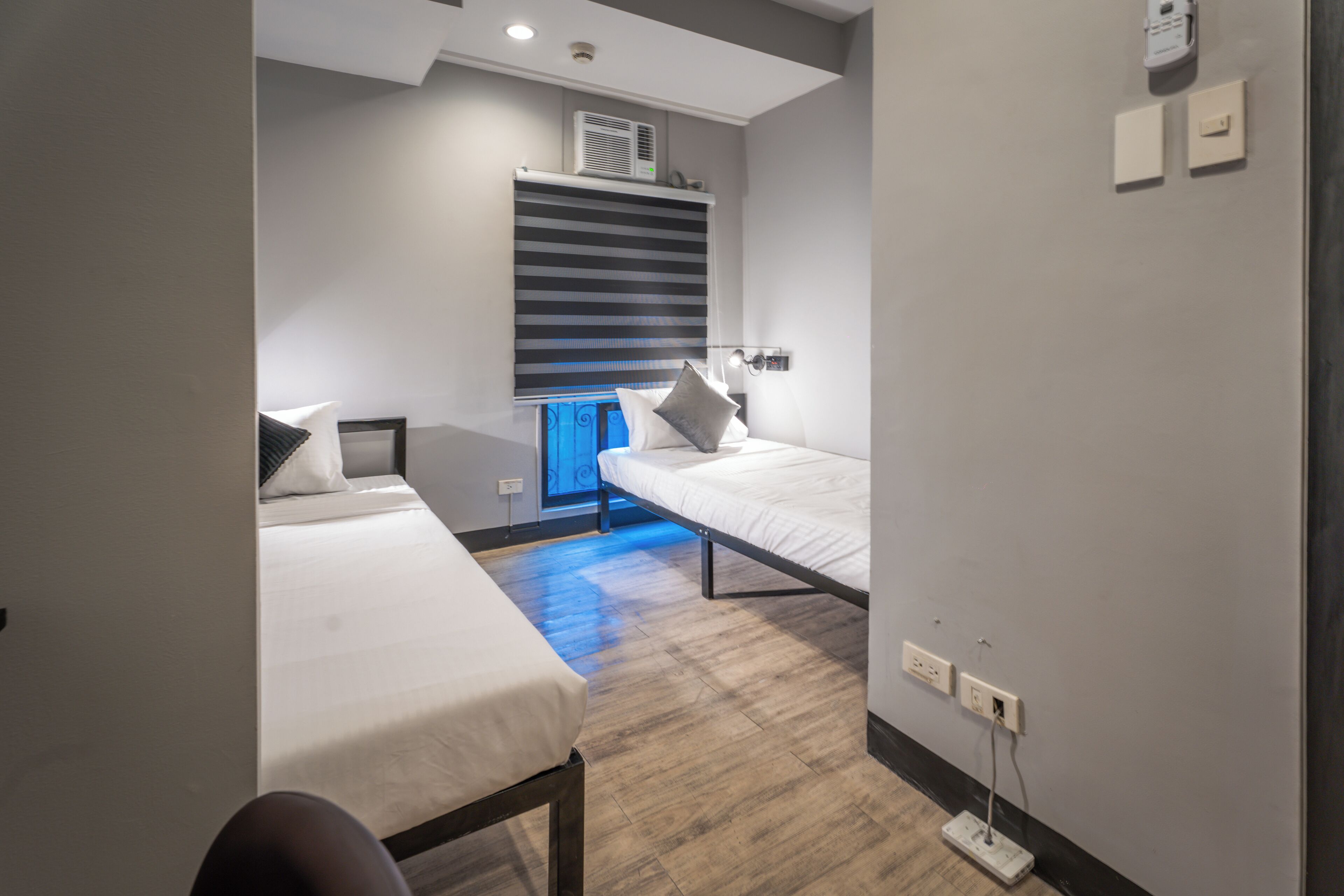 A modern hotel room with two single beds, a desk, and a window with blinds.