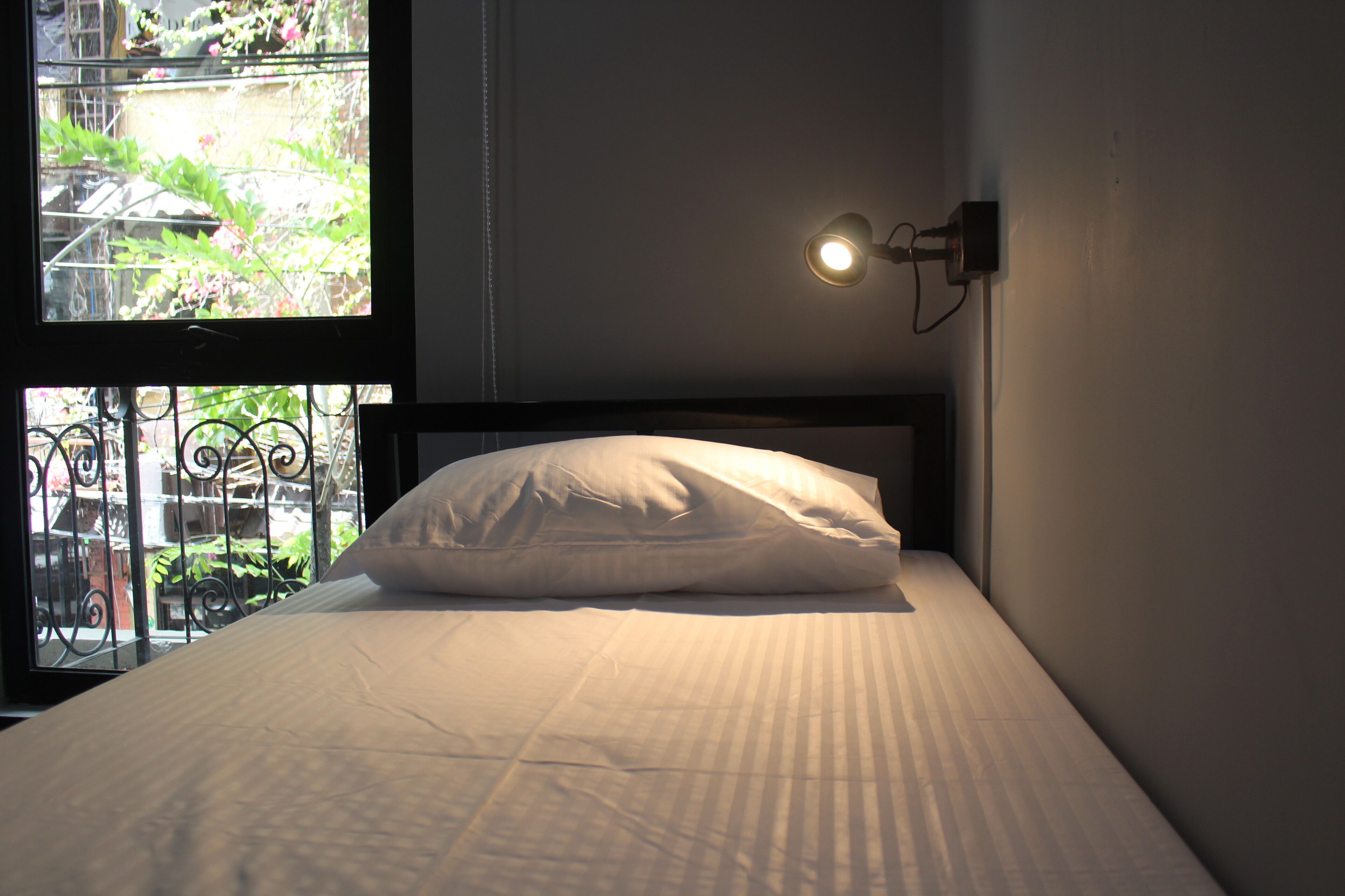 Un lit avec un oreiller blanc et une tête de lit, une lampe fixée au mur et une fenêtre donnant sur un paysage verdoyant, avec un balcon.