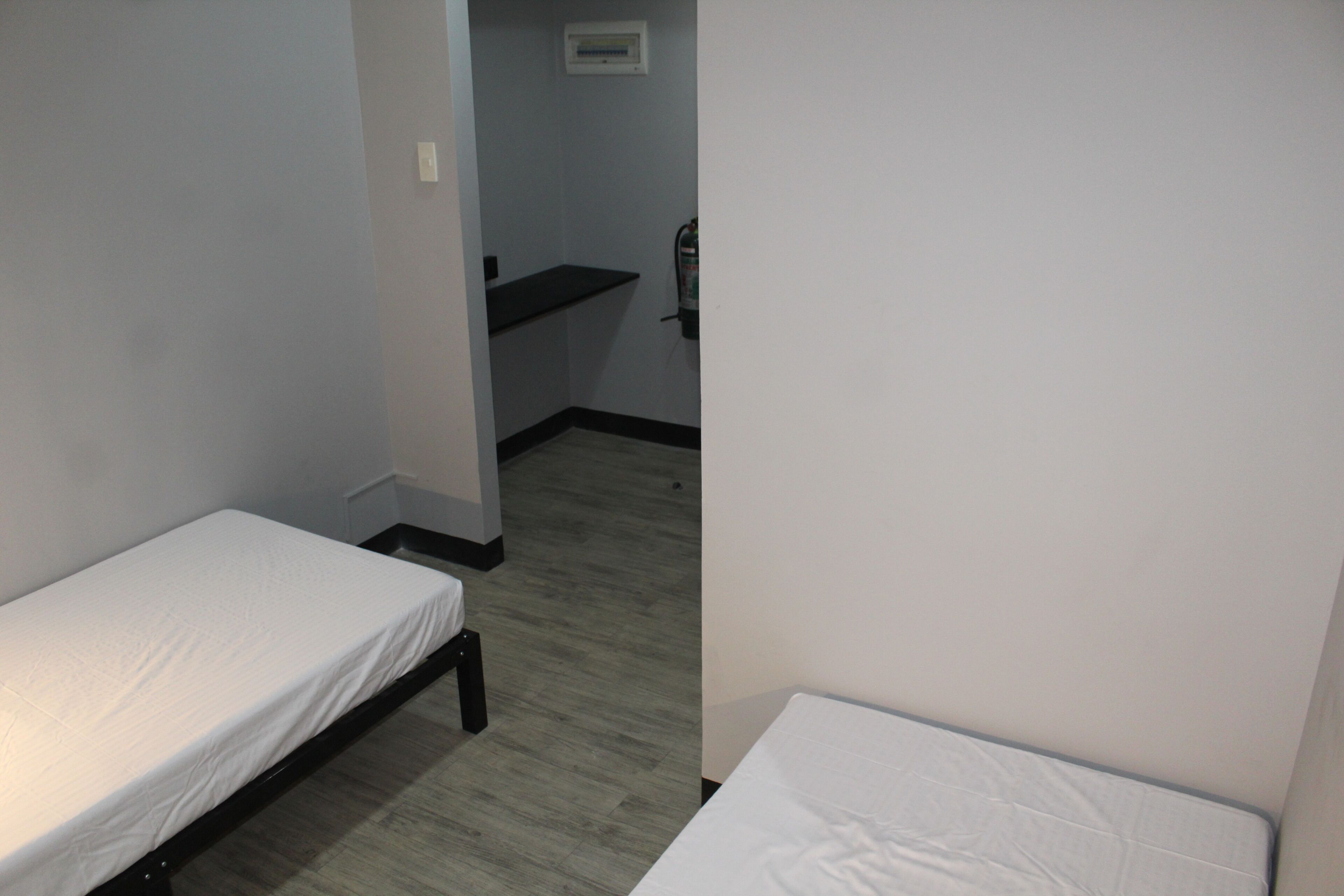 A small, narrow room with two single beds, a small shelf, and a wall-mounted light fixture.