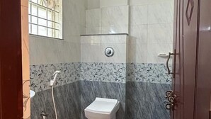 Une petite salle de bain carrelée, équipée de toilettes, d’une douche et d’un seau bleu.