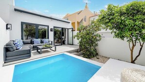 Ein moderner Poolbereich im Freien mit einer Sitzecke mit Sofa und Couchtisch, umgeben von einer weißen Mauer und einem Baum.