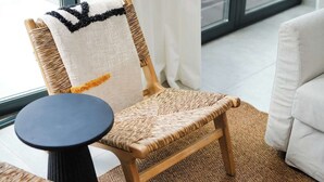 Ein Korbstuhl mit gewebter Sitzfläche und Lehne, ein runder schwarzer Tisch und ein weißes Sofa in einem Zimmer mit einem großen Fenster.