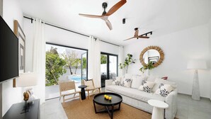 Ein modernes Wohnzimmer mit einer Couch, einem Couchtisch und Sesseln, einem Fernseher, Deckenventilatoren und Blick auf einen Pool.