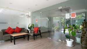 A modern reception area with a glass wall, a sofa with red cushions, a wooden table, and potted plants. - Hotel Royal Galaxy (Gurugram)