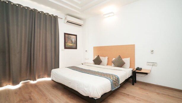 A hotel room with a wooden floor, a bed with white bedding, a nightstand with a phone, and a wall-mounted air conditioner. - Hotel Royal Galaxy (Gurugram)