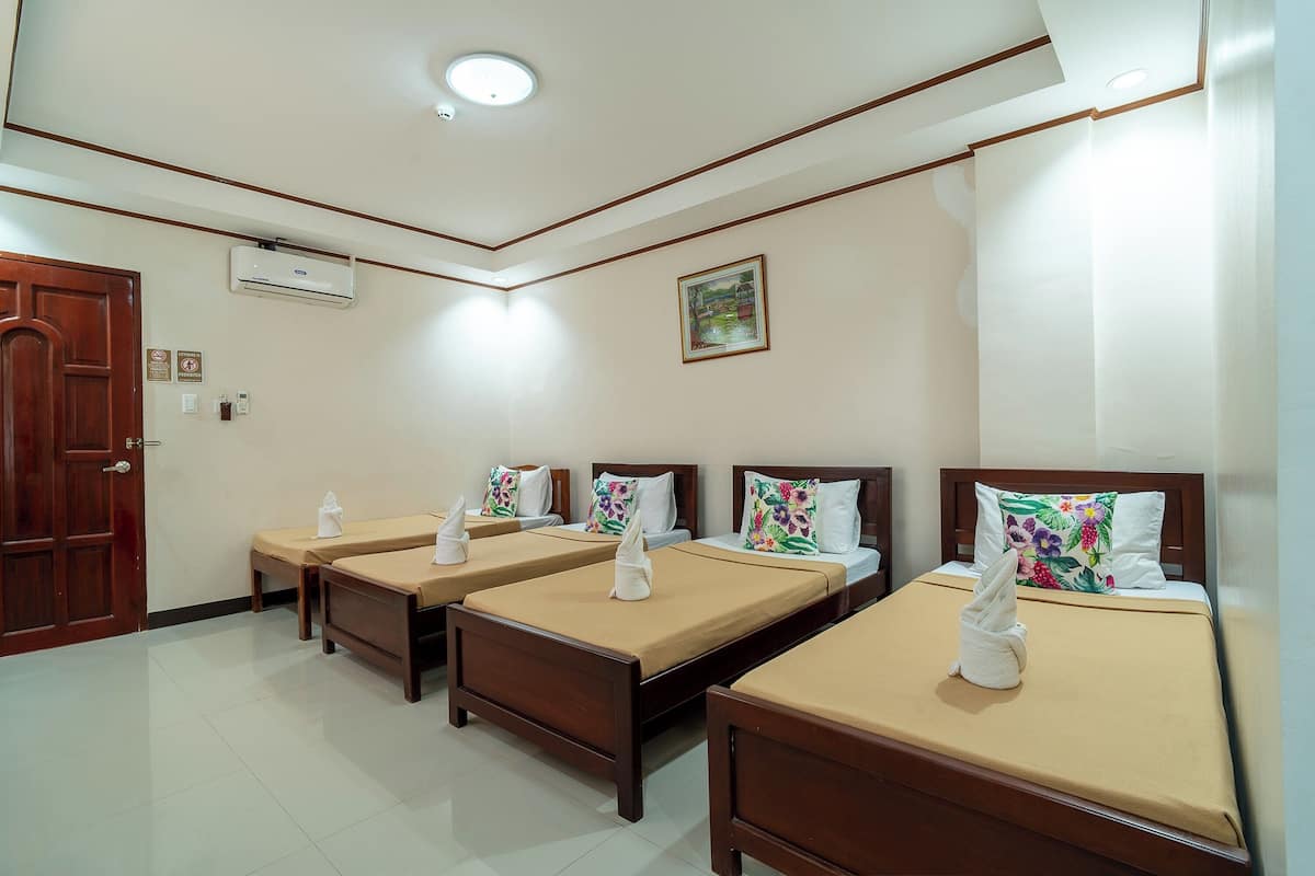 A hotel room with four single beds, each with a brown headboard, beige bedsheets, and a white towel on the pillows. There is a wooden door, an air conditioning unit on the wall, and a framed picture above the beds.