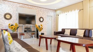 A modern living room with a brown sofa, a wooden coffee table, and a flat-screen TV. Sunflowers are placed on the coffee table, and there is a bookshelf with decorative items. - Dinah's Tourist Inn Puerto Princesa (Puerto Princesa)