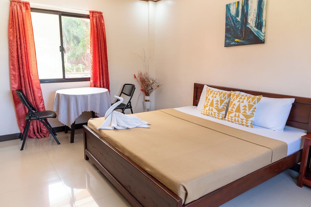 A neatly made bed with a brown bedspread, a small round table with a white tablecloth, a chair, and a vase with dried branches.