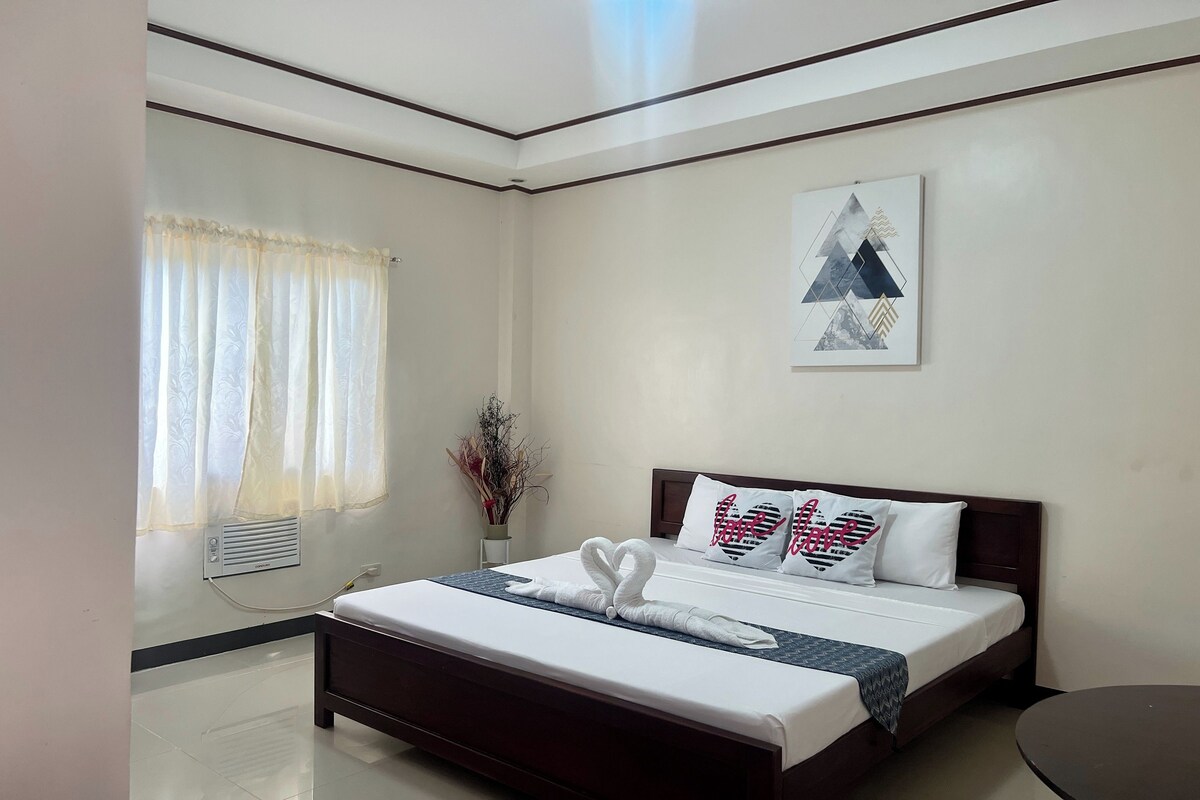 Quarto de hotel com uma cama bem arrumada, uma mesa de cabeceira, uma obra de arte na parede e uma janela com cortinas.