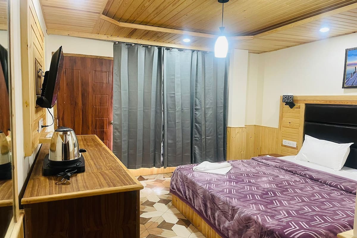 Une chambre d’hôtel avec un plafond en bois, un lit recouvert d’une courtepointe à motifs, une table de chevet avec une bouilloire, et une vue sur l’extérieur par une fenêtre munie de rideaux.