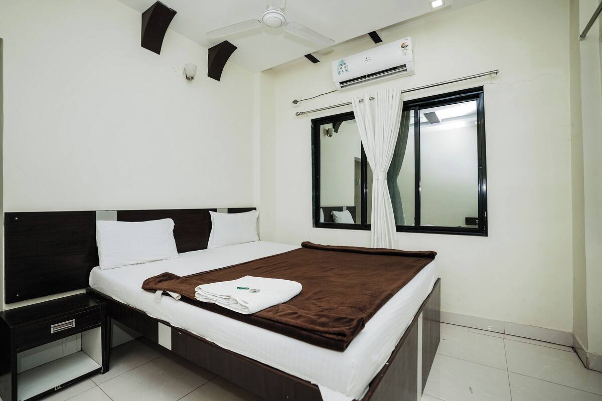 A neatly made bed with a brown blanket, a bedside table, a nightstand, a ceiling fan, an air conditioning unit, and a window with curtains.