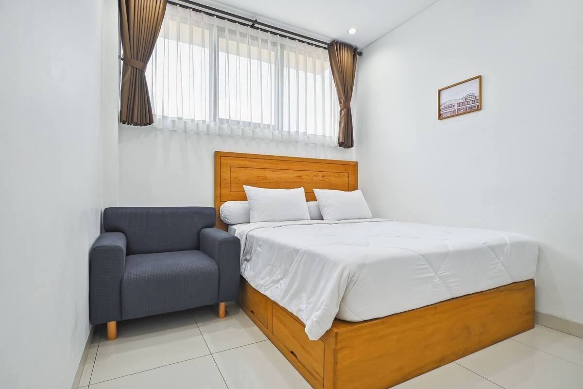 A bedroom with a wooden bed, white bedding, a grey sofa, and sheer curtains.