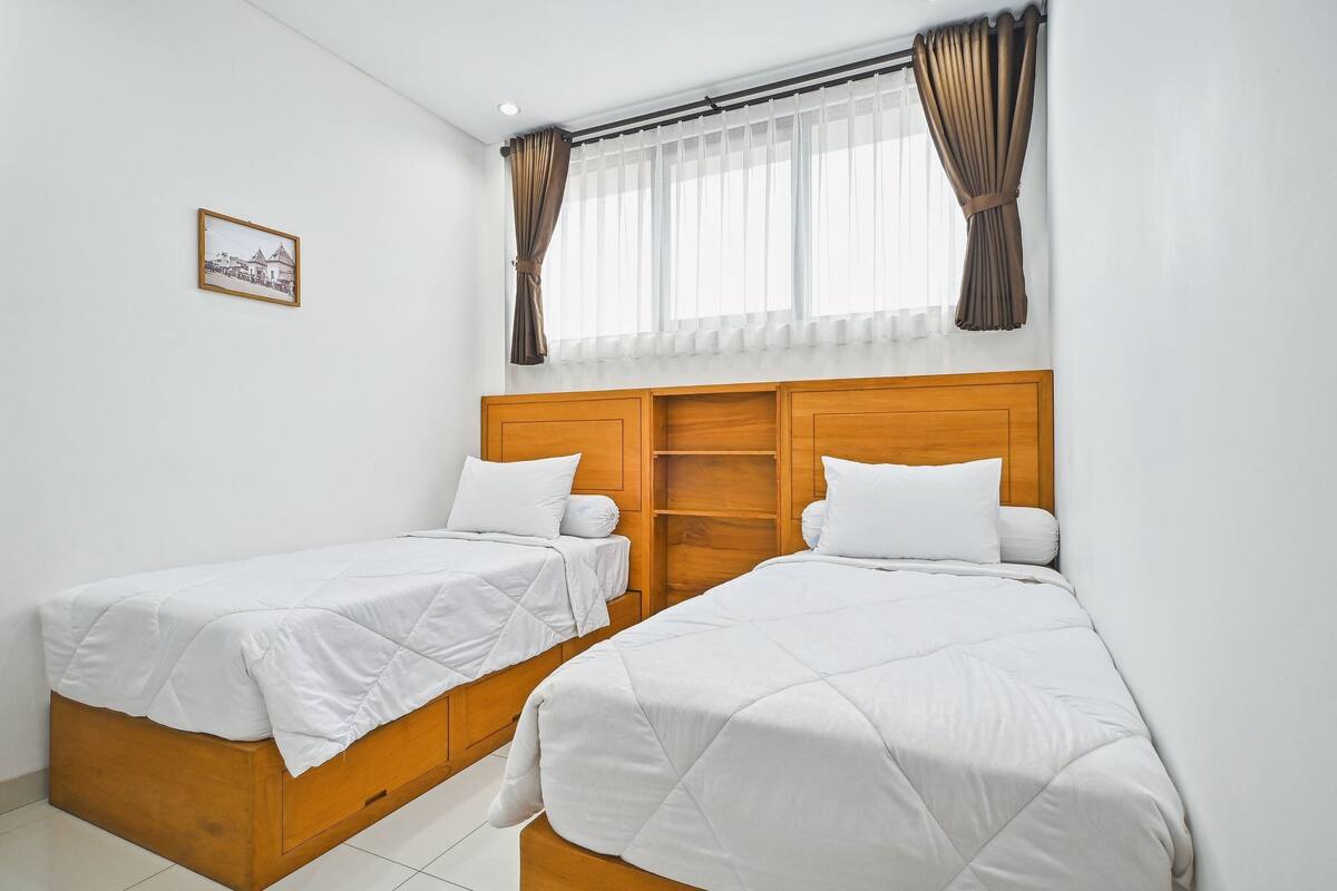 A room with two single beds, wooden headboards, a small picture on the wall, and a window with curtains.