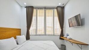 A hotel room with a bed, a wooden headboard, a TV mounted on the wall, a small table with toiletries, and a window with curtains. - Bangreng Inn (Bandung)