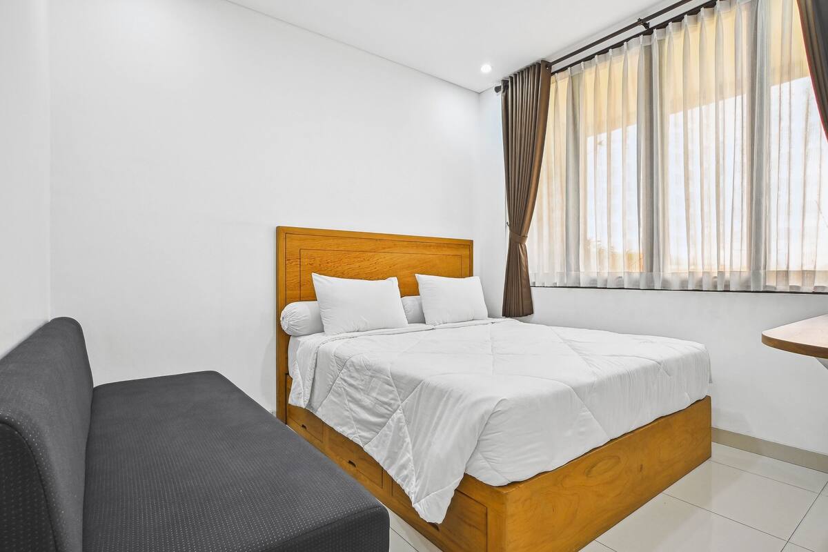 A bedroom with a wooden bed, white bedding, a grey sofa, and a window with curtains.