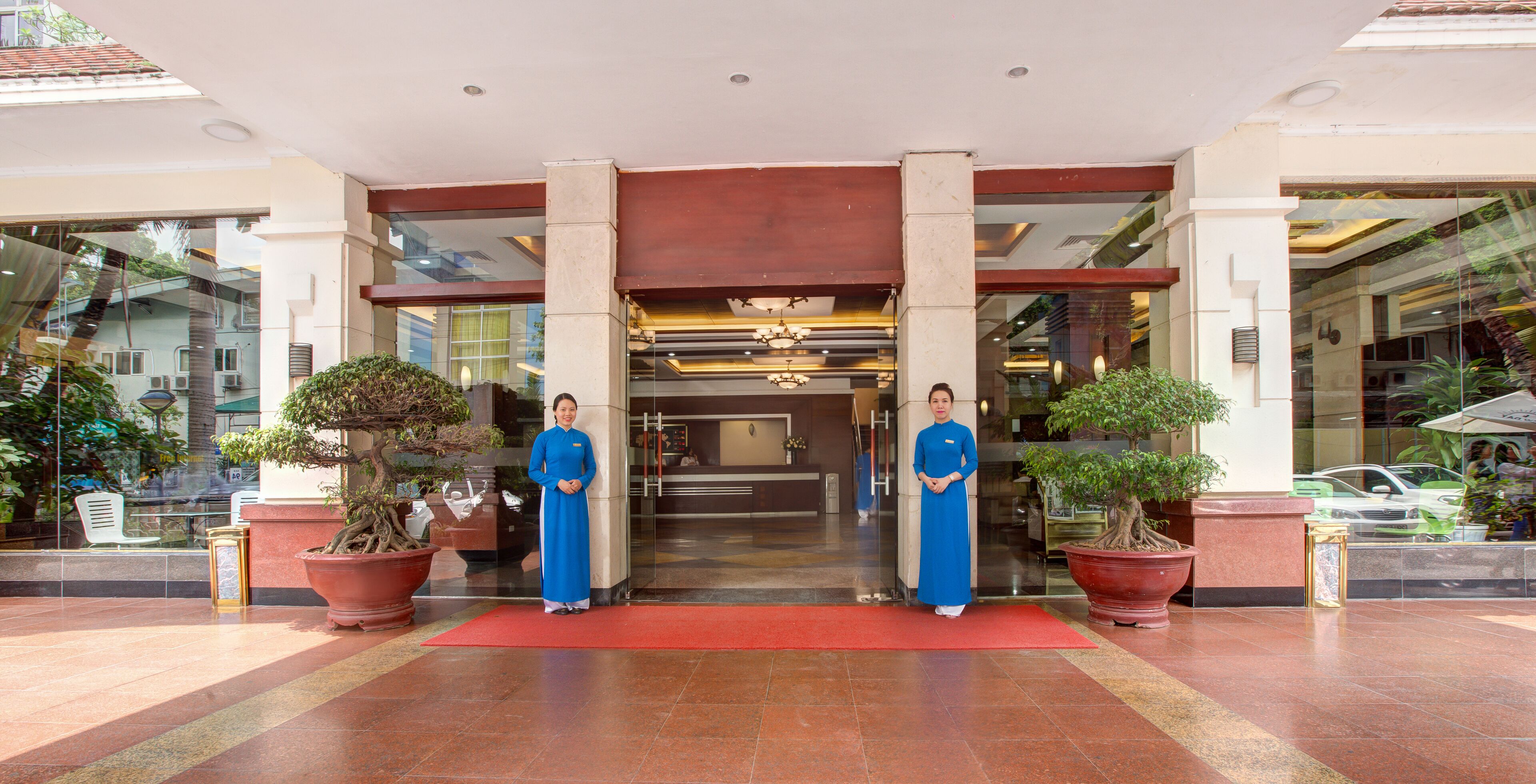 A hotel entrance with two receptionists, a red carpet, and potted plants.