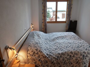 Ein Schlafzimmer mit Bett, Nachttisch, einem Fenster mit Vorhängen und Blick ins Freie.