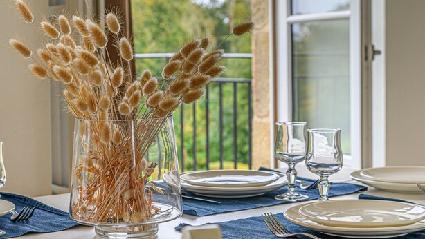 A dining table with a clear glass vase containing dried plants, set with white plates, silverware, and wine glasses. - Château De Kergonano by Interhome (Baden)