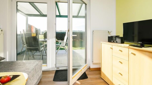 Una stanza con una porta scorrevole in vetro che dà su un patio, una televisione su un mobile in legno e un tavolino con una ciotola di frutta.
